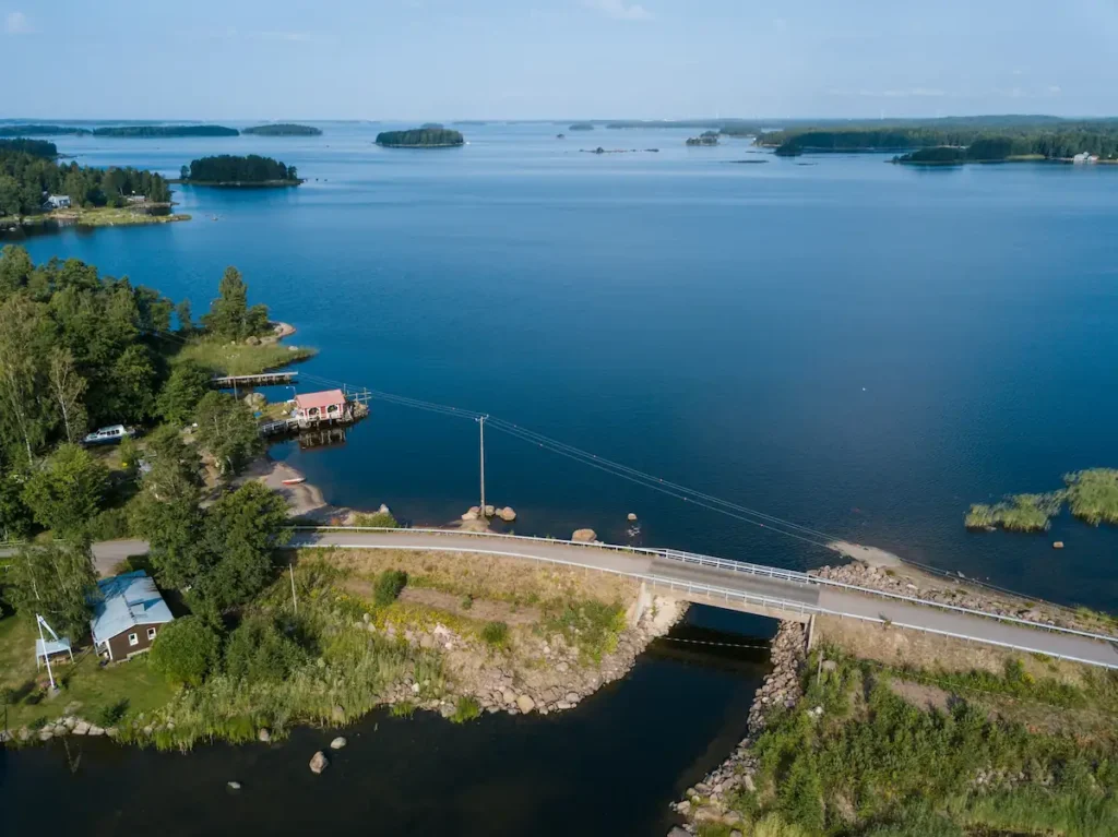 Ilmakuva Saariston pieneltä rengastieltä, jossa silta kulkee pienen salmen yli meren rannalla, taustalla punainen venevaja, mökki ja saaristo kesämaisemassa.