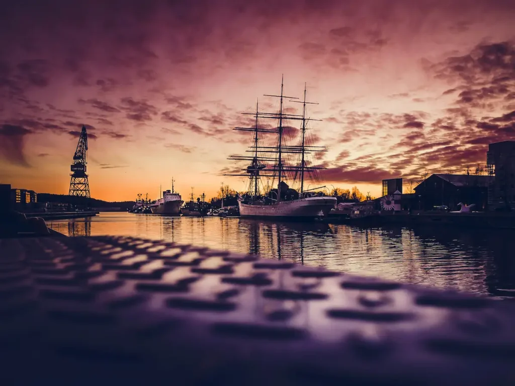 Kolmimastoinen purjelaiva ankkuroituna Turun satamassa auringonlaskun aikaan, taustalla historialliset telakat ja meri.